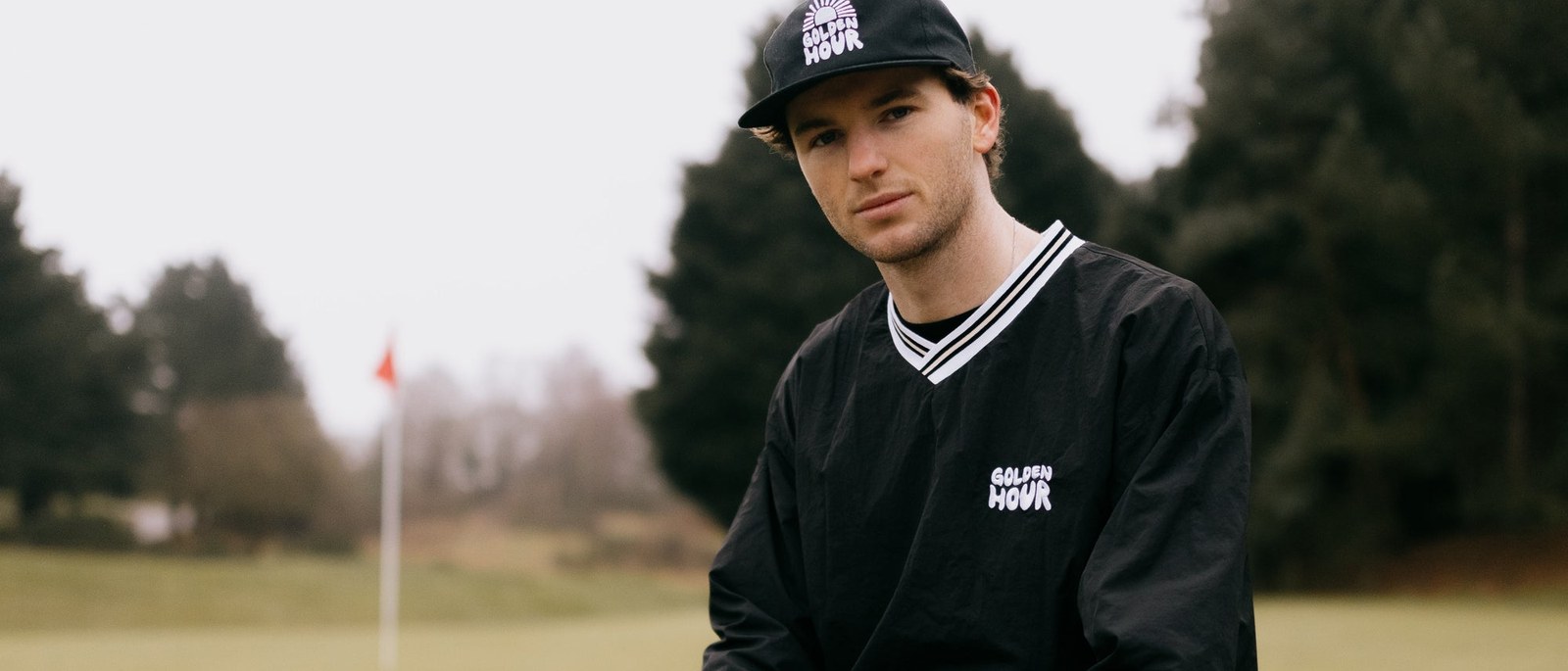 A golfer in a black Golden Hour windbreaker and cap sitting on the course with a flag behind him