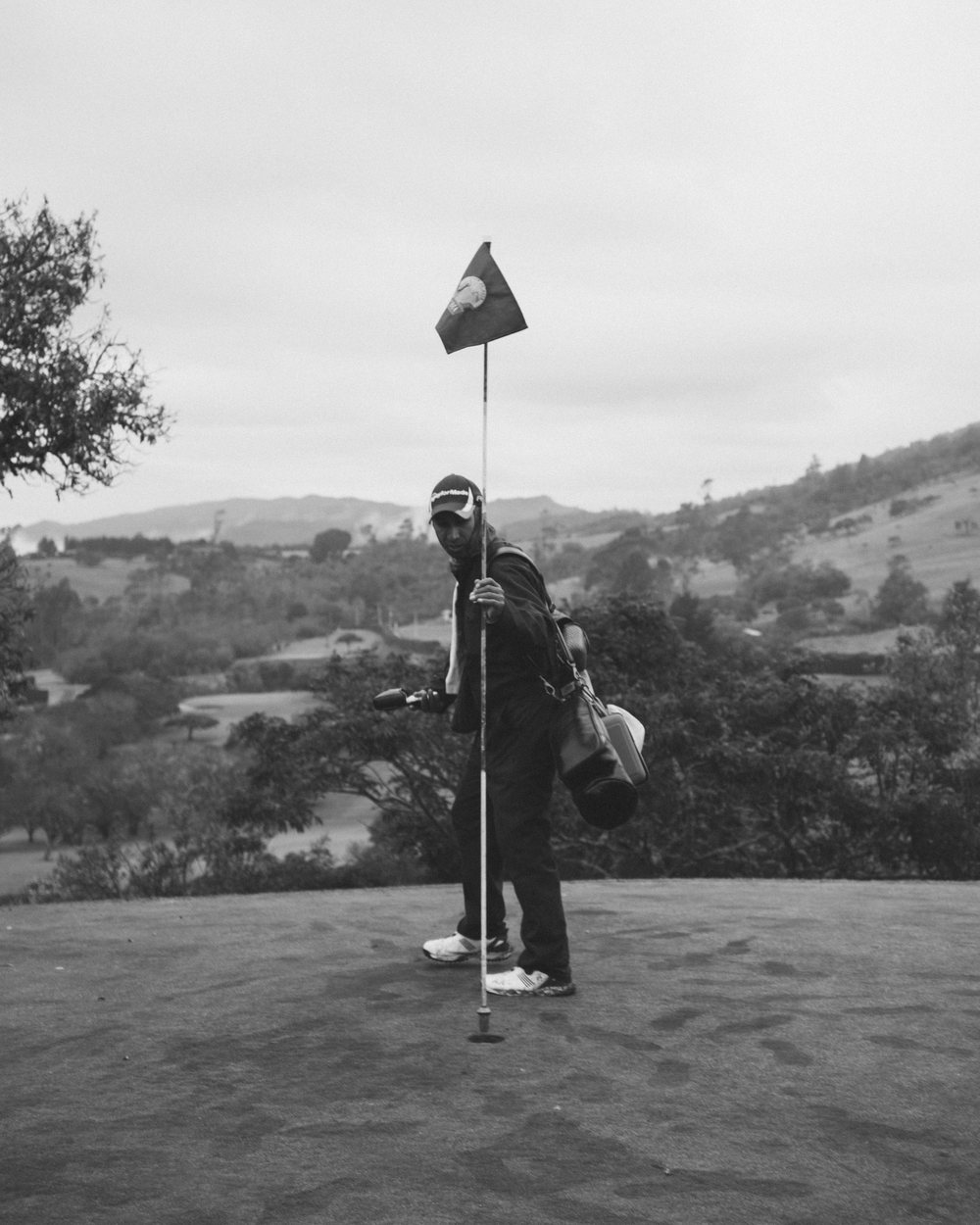 A golfer holding the flag on a green with hills behind, in black and white