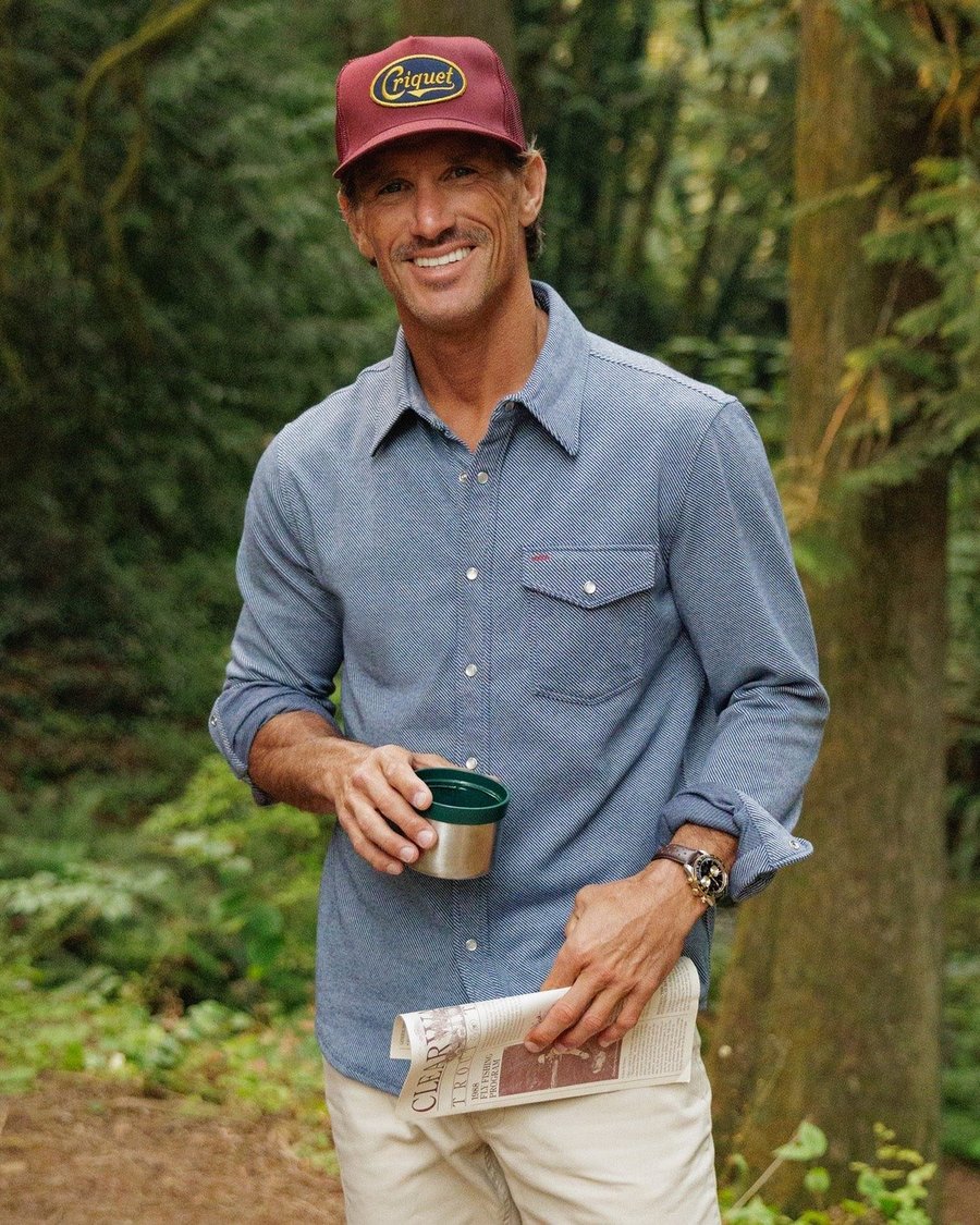 A smiling man in a chambray Criquet shirt and cap holding a cup