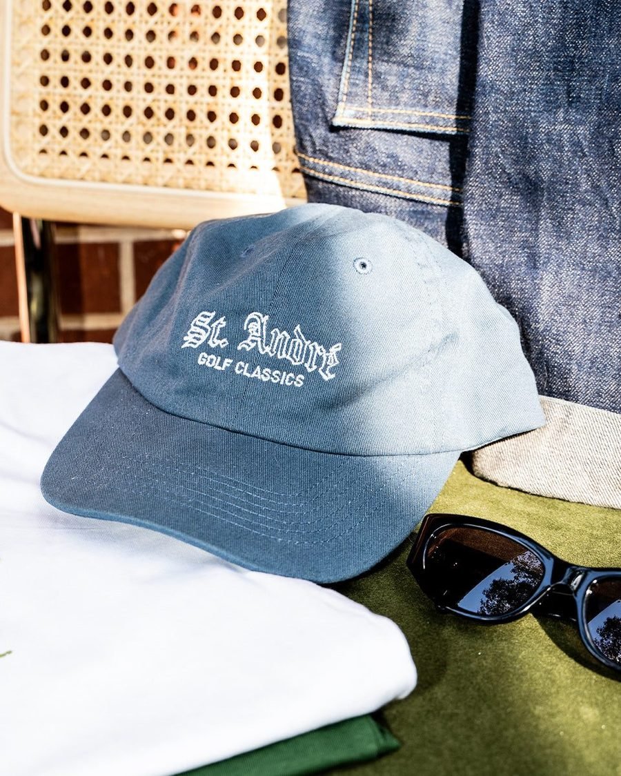 A light blue St Andr&eacute; cap on a white tee beside sunglasses and denim