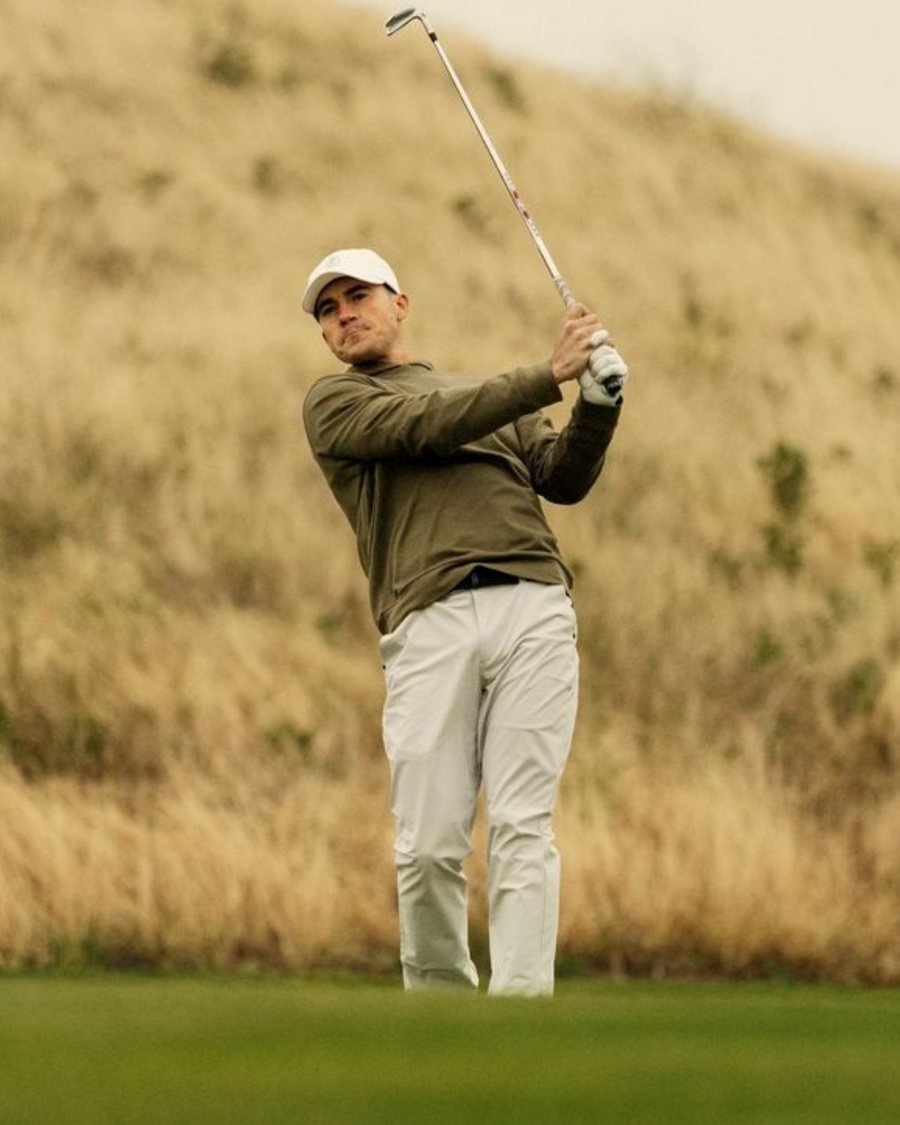 A golfer in an olive Ashworth layer finishing his swing beside a grassy dune