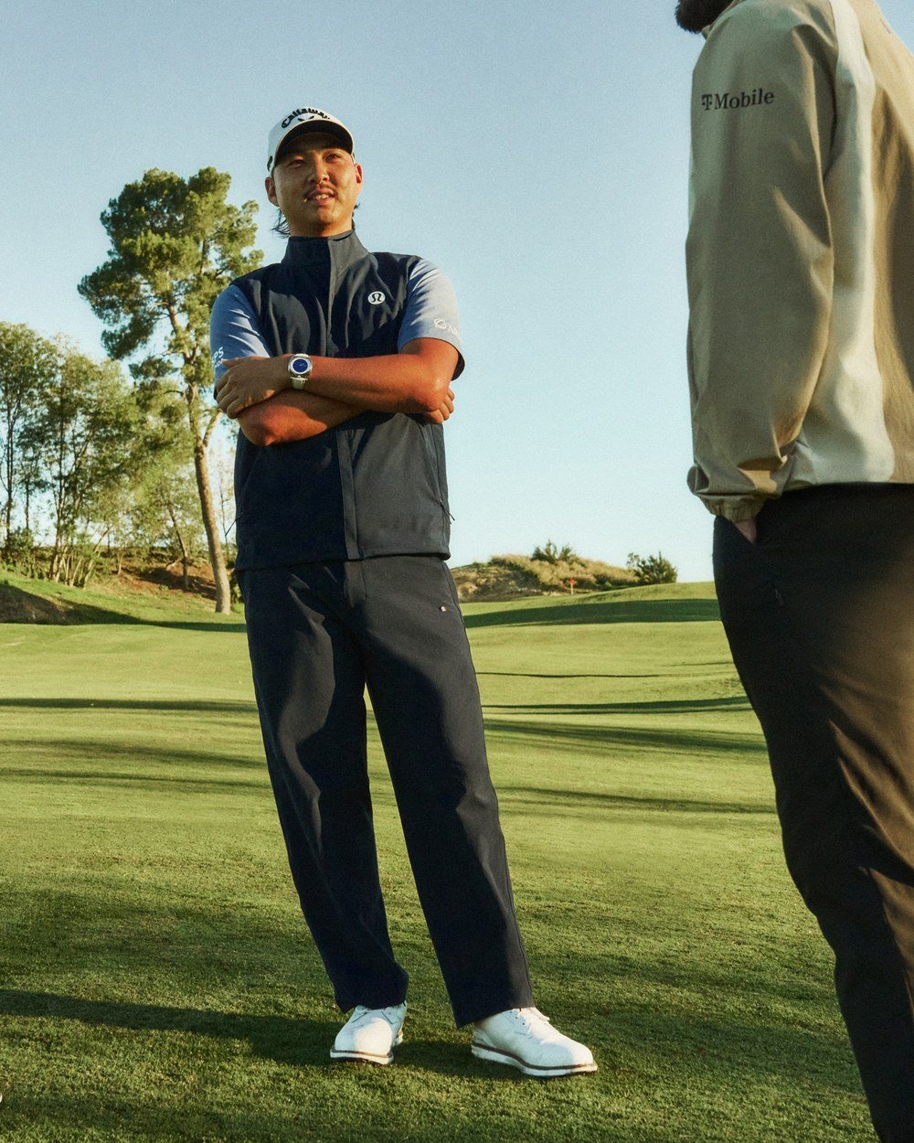 Min Woo Lee in a navy vest and dark trousers on a golf course
