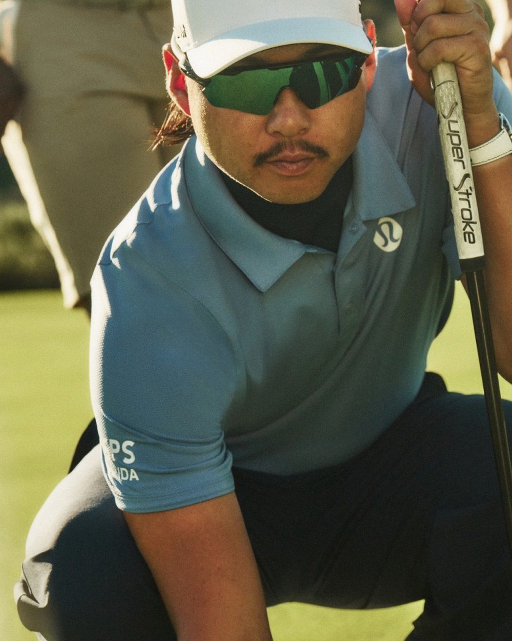 Min Woo Lee crouched reading a putt in a blue polo and sunglasses