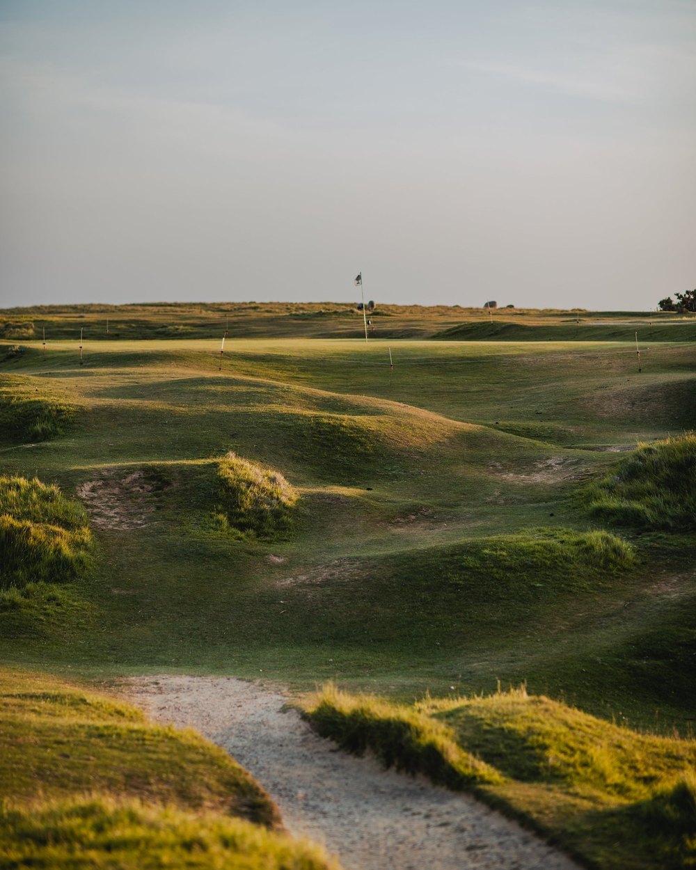 A sandy path winding between grassy dunes toward a green on an English hilltop course