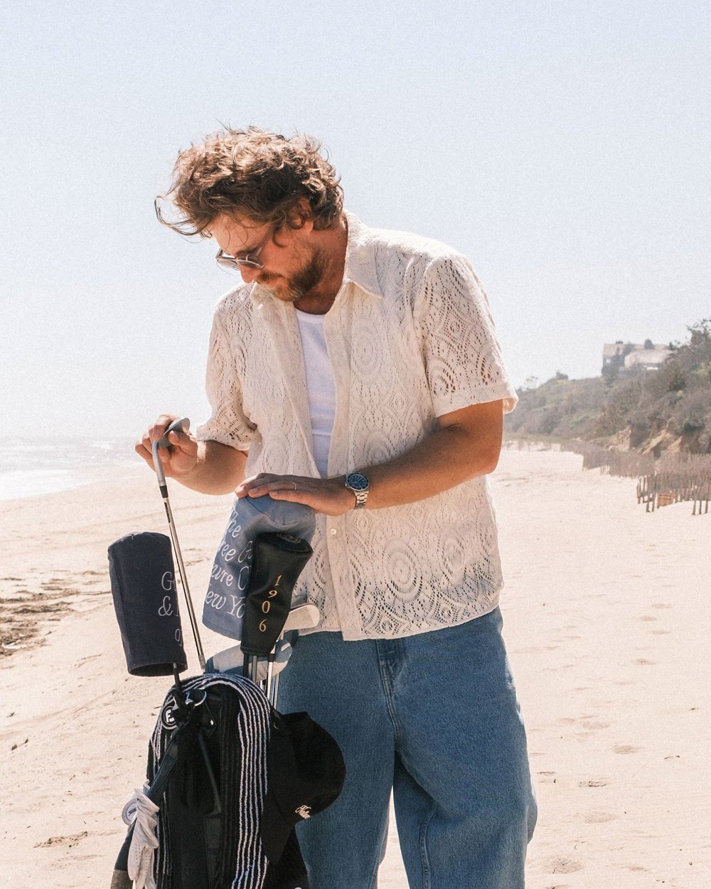 A man in a lace short-sleeve shirt and jeans checking a golf bag on a beach
