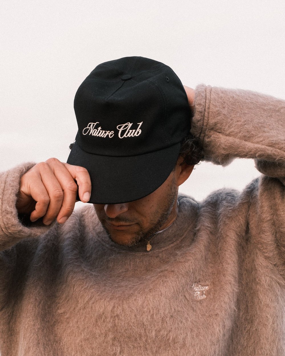 A man in a fuzzy sweater adjusting a black Nature Club cap
