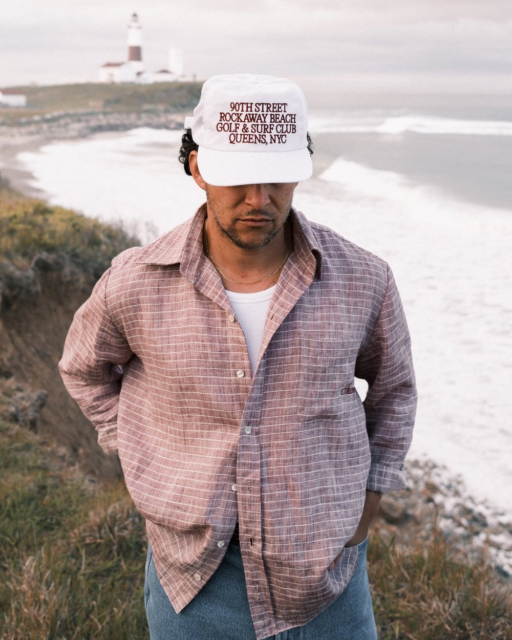 A man in a pink check linen shirt standing on a cliff above the ocean with a lighthouse behind