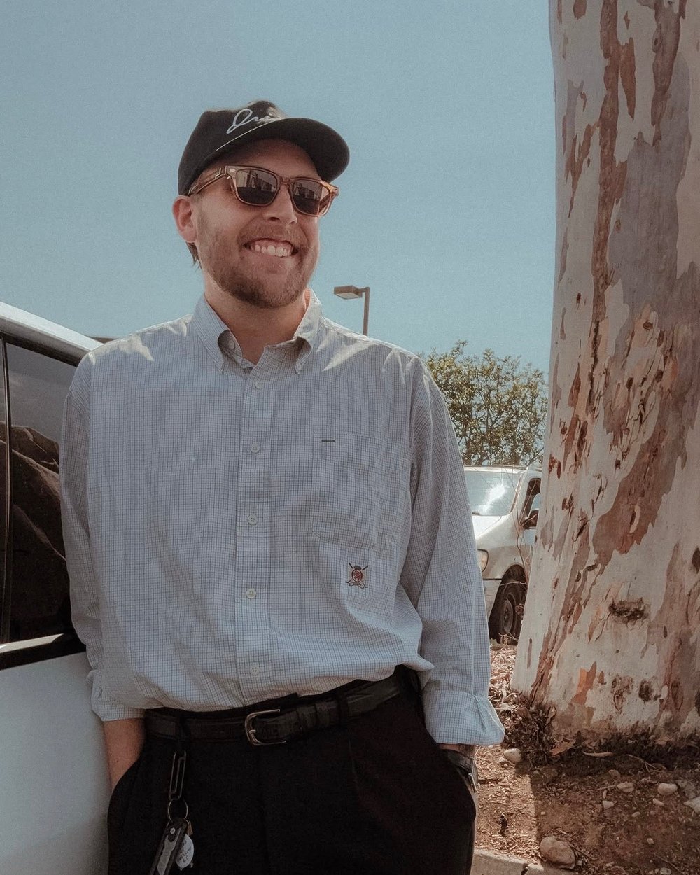 A man in a black cap and sunglasses leaning against a car beside a eucalyptus tree