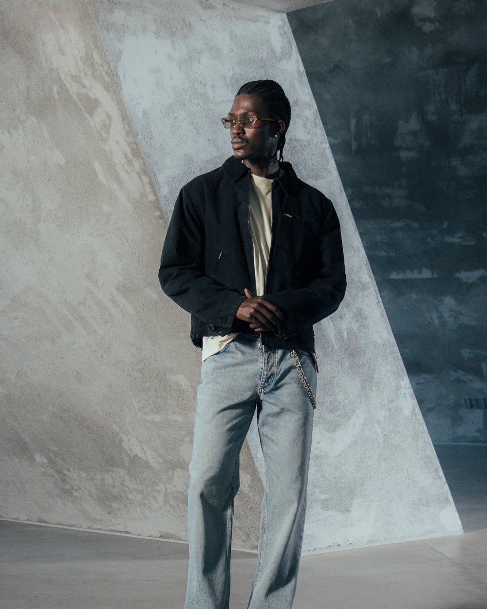 A man in a black jacket and light jeans against a concrete wall