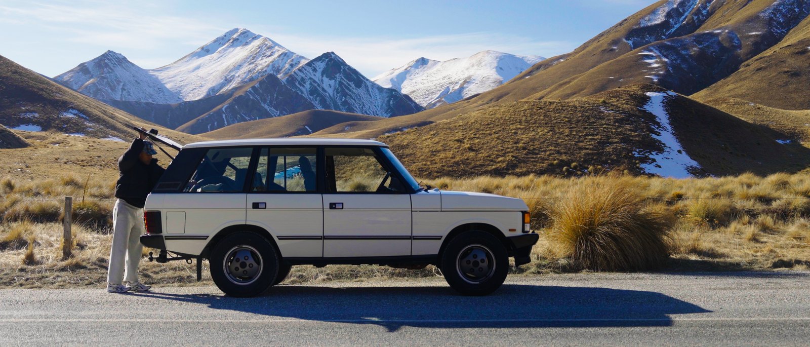 A vintage white 4x4 parked on a road through a mountain valley, snow on the peaks and tussock grass in the foreground