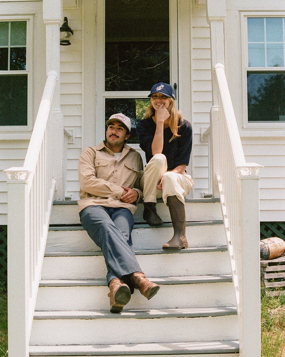 Two people in Communal hats sitting on the front steps of a white clapboard house