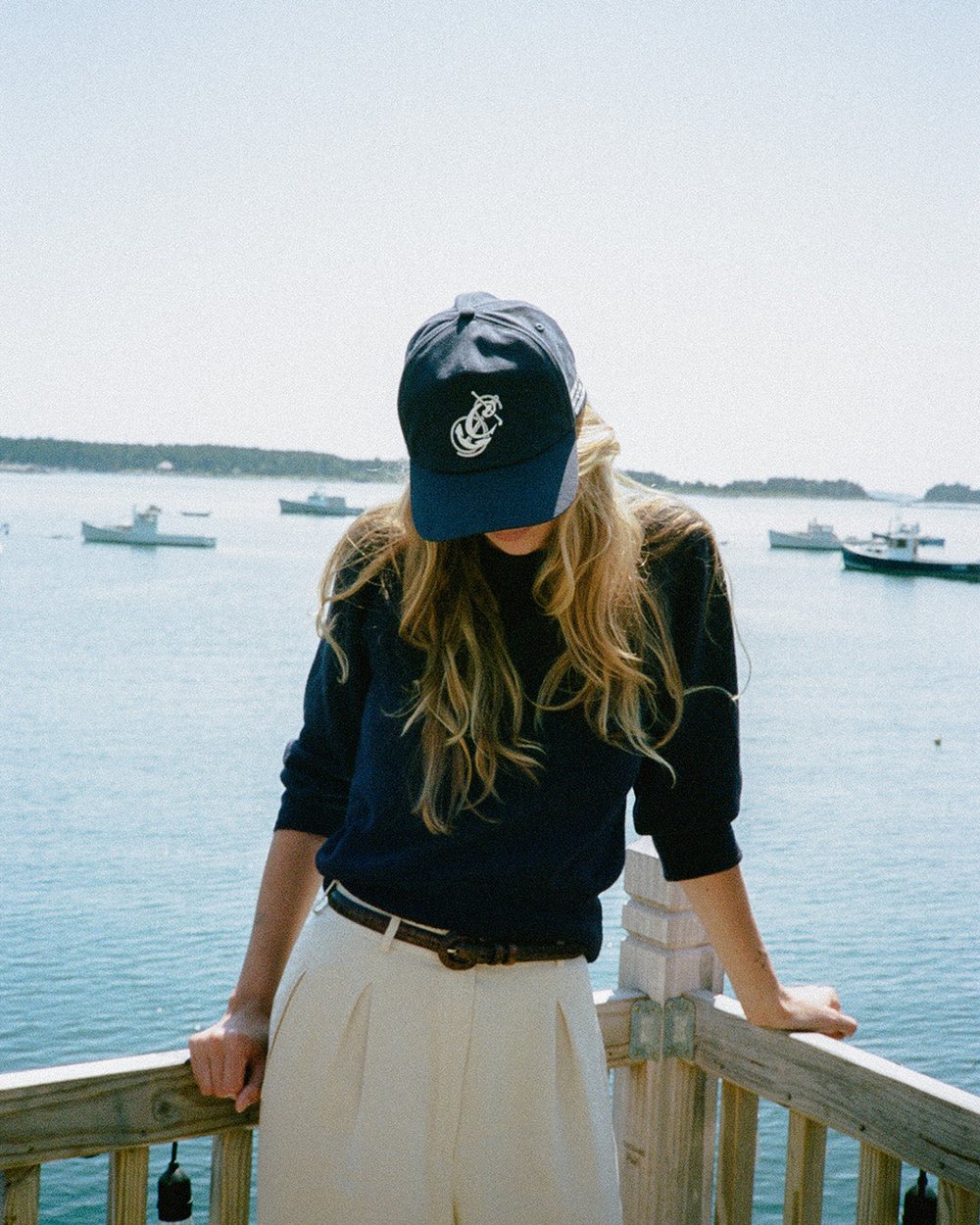 A woman in a navy Communal anchor hat leaning on a railing over a harbor