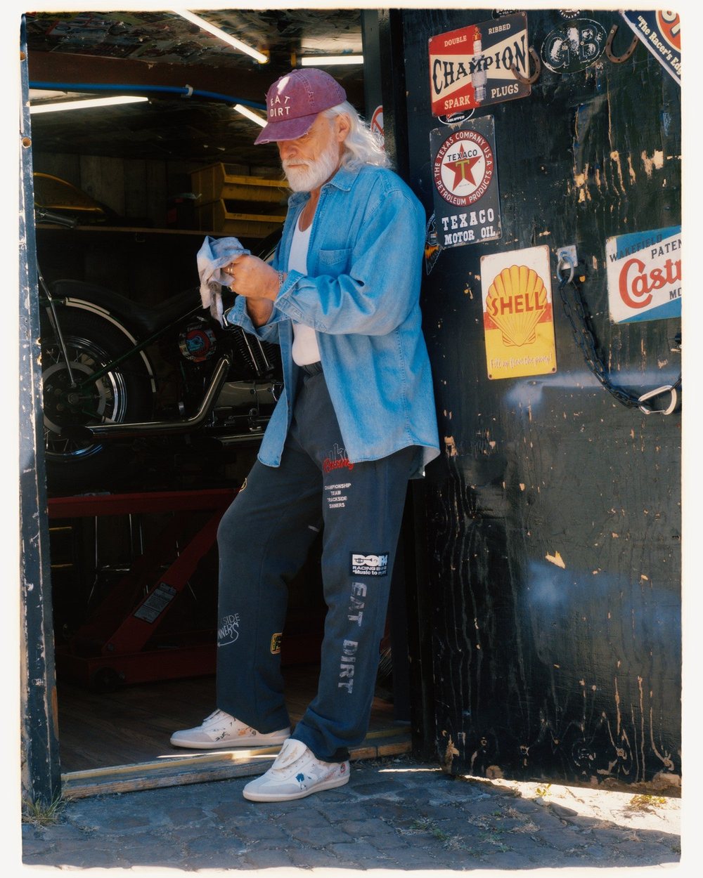 A man in an Eat Dirt cap and denim shirt standing in a garage doorway covered in old oil signs