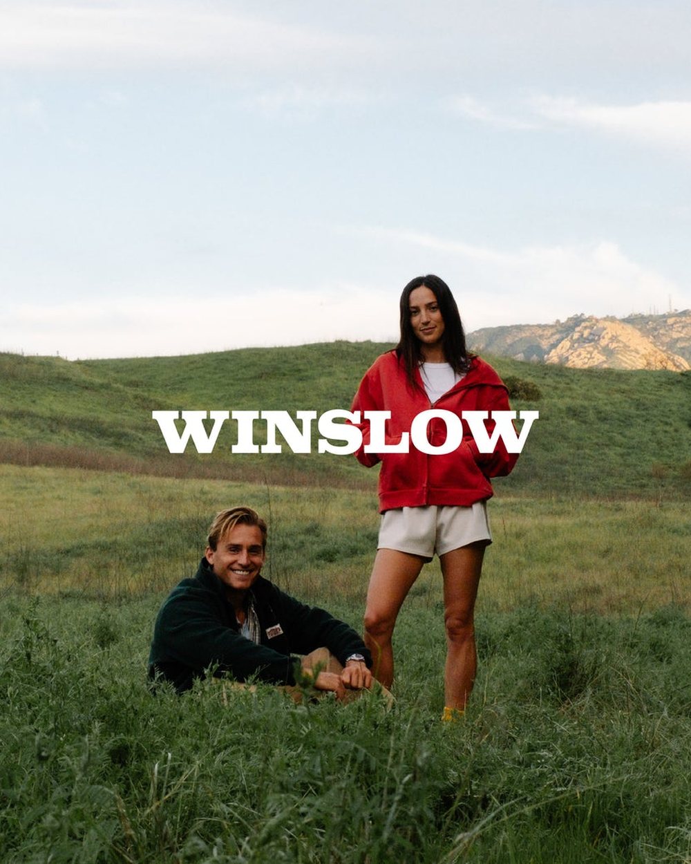 Two people in Winslow sweatshirts in a green field below California hills