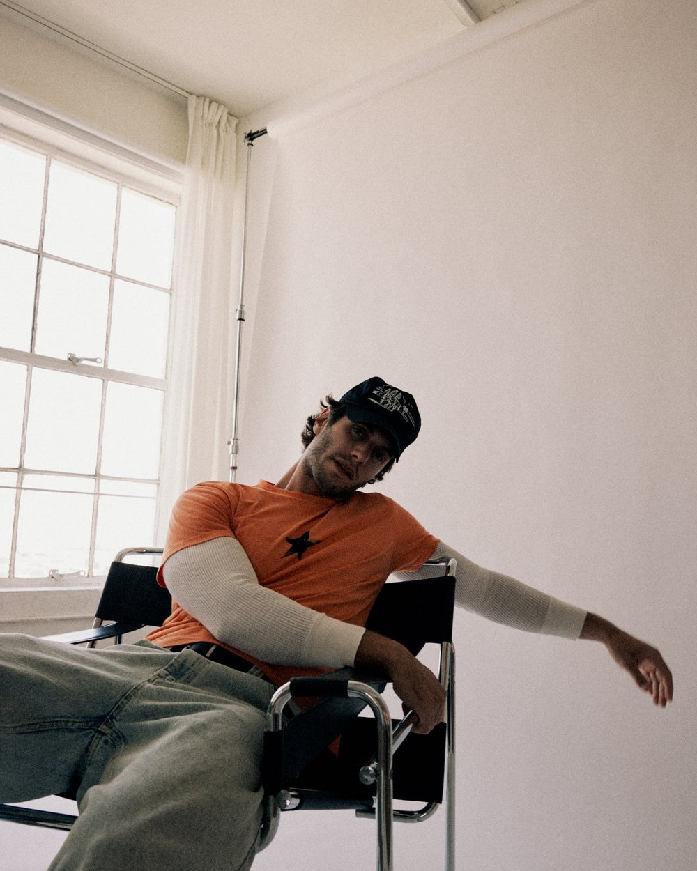 A man in an orange top and dark cap slouched in a chair in a bright studio