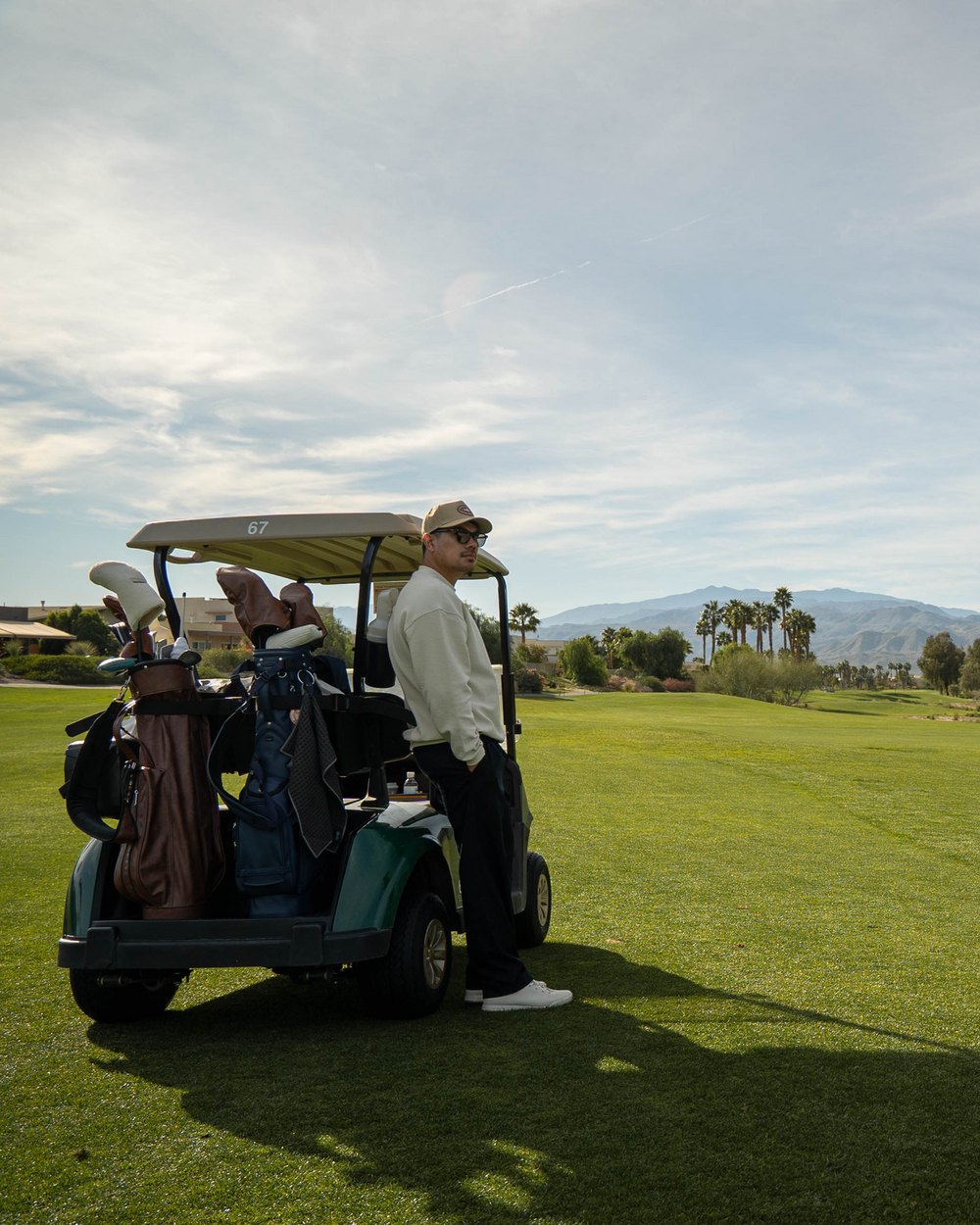 A golfer in a cream sweater and cap leaning on a golf cart on a desert fairway