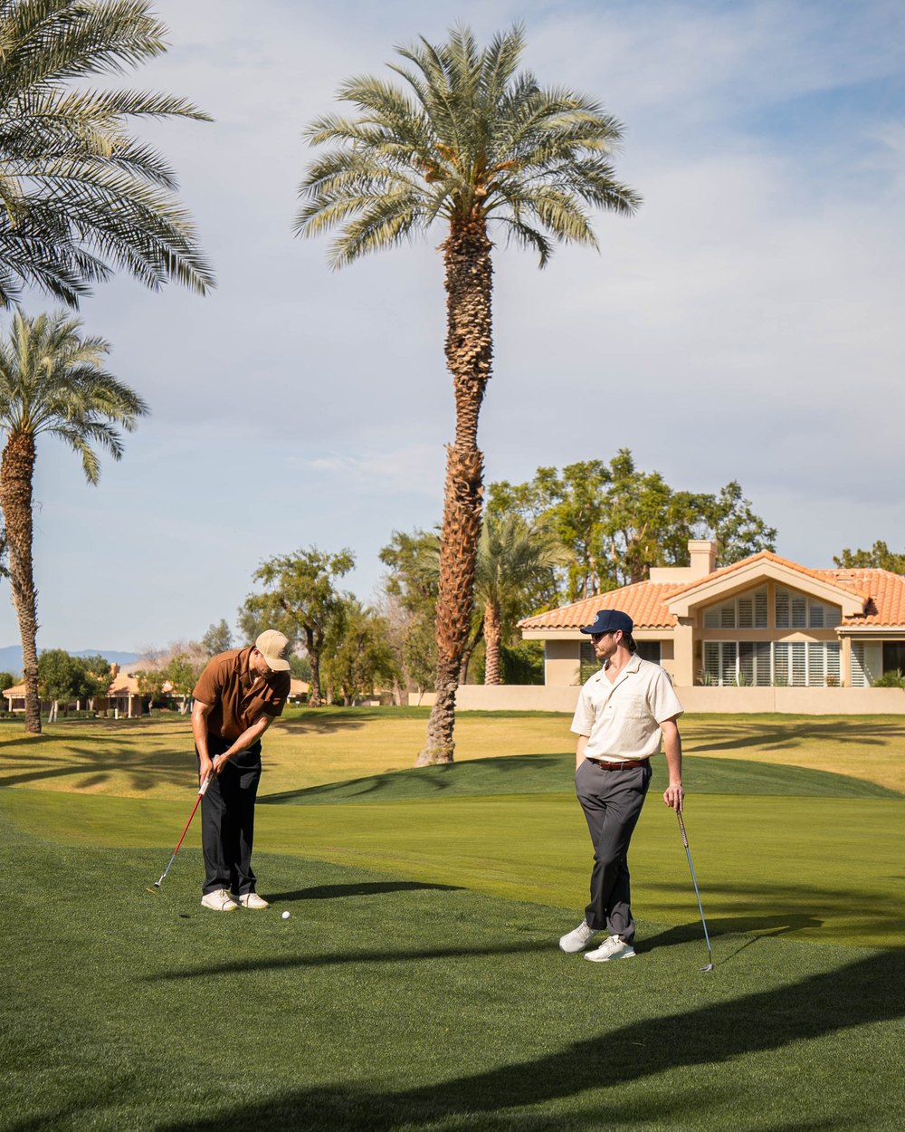 Two golfers on a green under tall palms, one putting