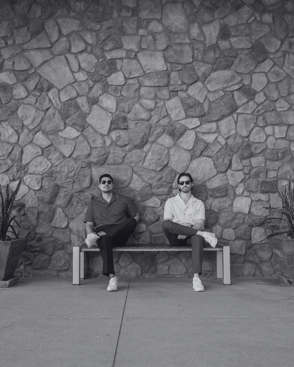 Two men in Howie Benson polos on a bench against a stone wall, in black and white