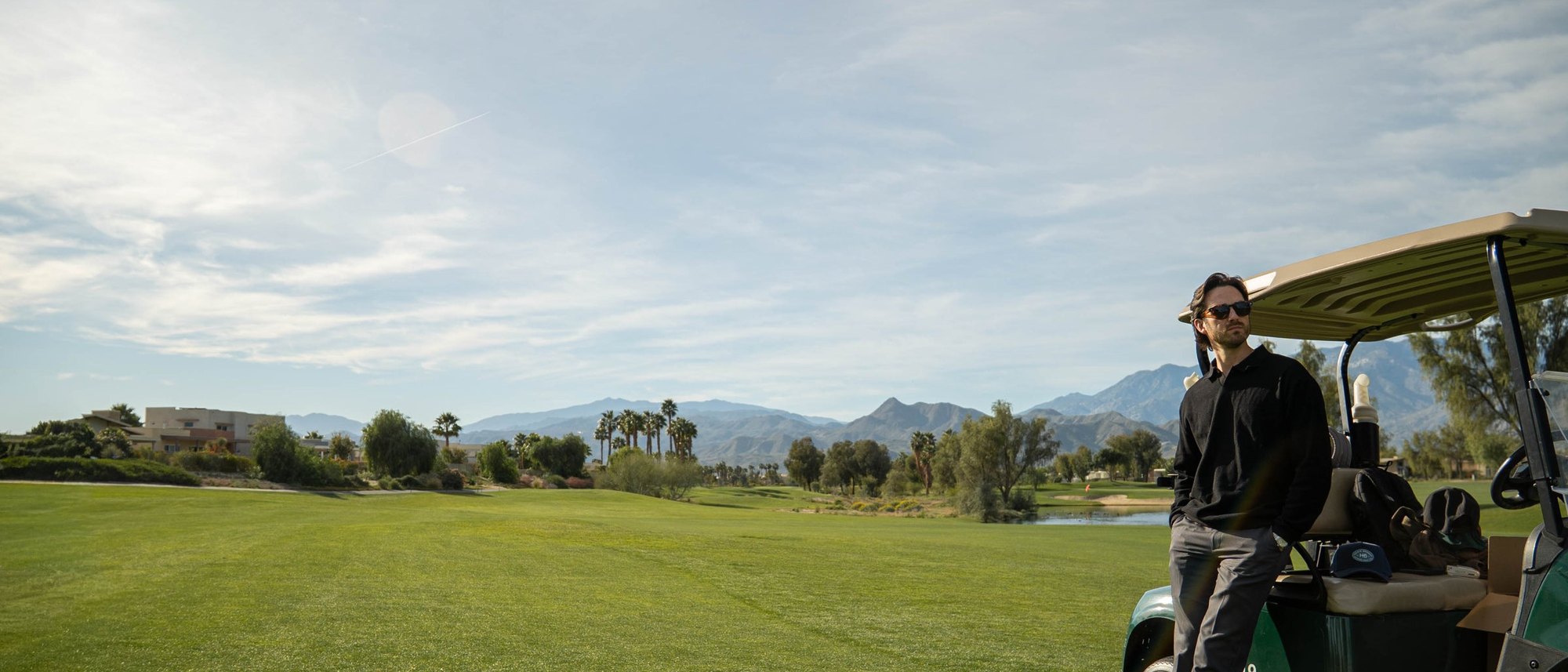 A golfer in a black Howie Benson pullover and sunglasses leaning on a golf cart on a desert course below the mountains