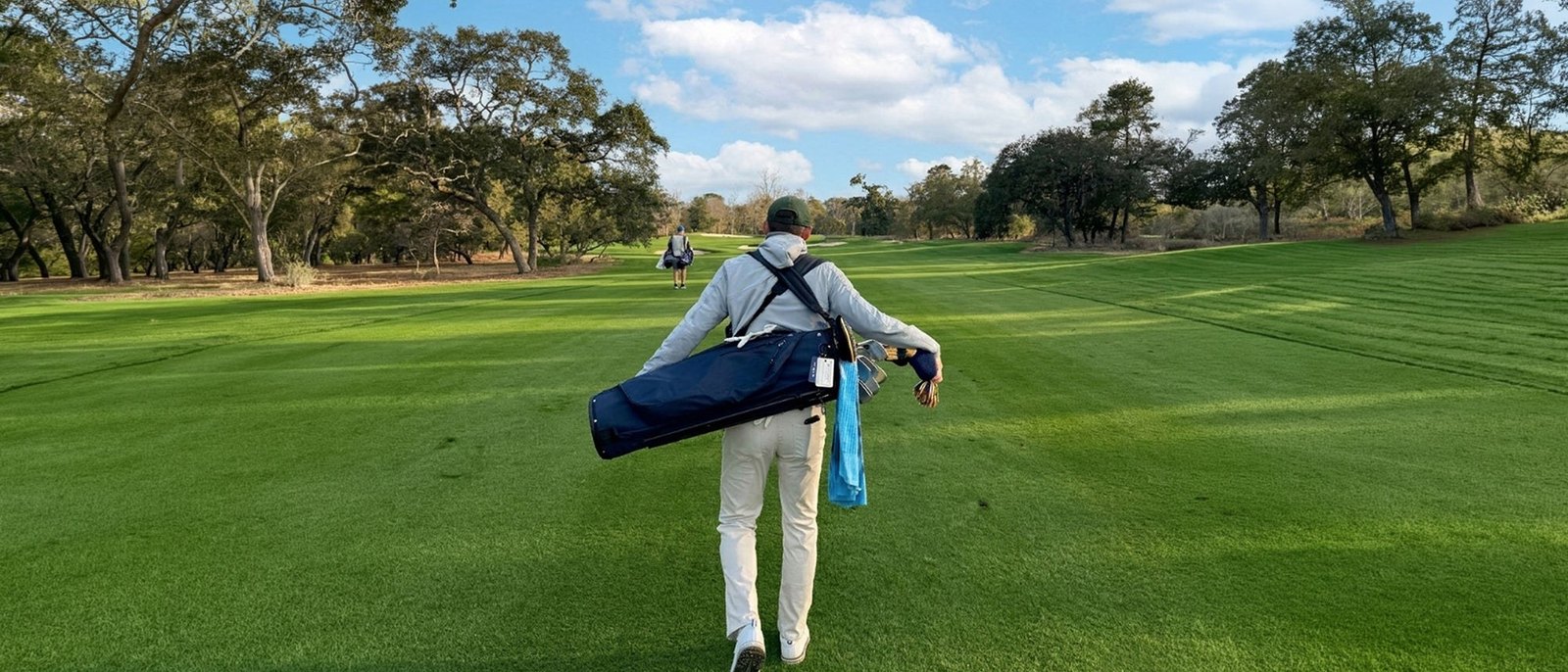 A golfer walking down a fairway under oak trees with a navy Hudson Sutler carry bag on his back