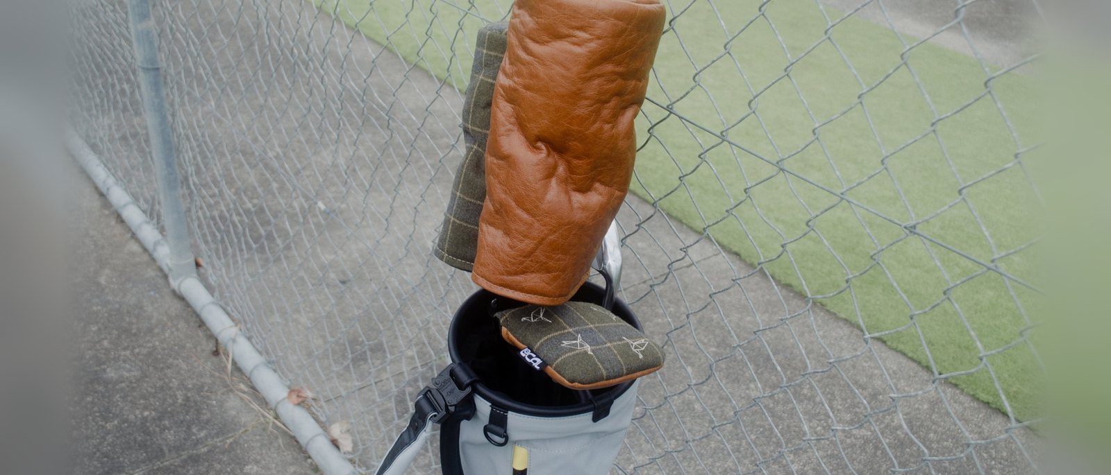 A tan leather Local GC headcover on a driver in a golf bag beside a chain-link fence at the range