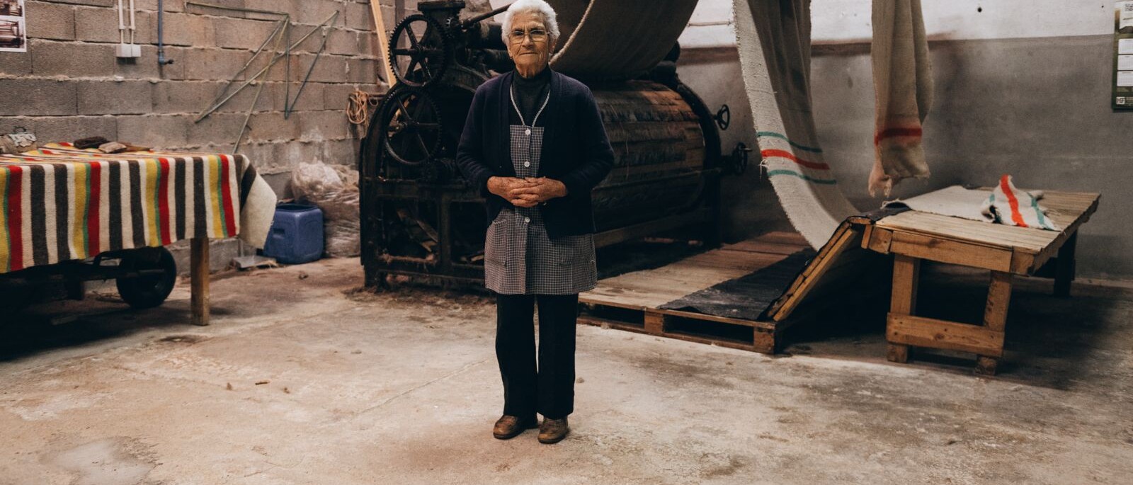 A weaver standing at a manual wooden loom in the Portuguese wool mill that makes Mogshade’s headcover textiles, striped blankets stacked on the bench beside her
