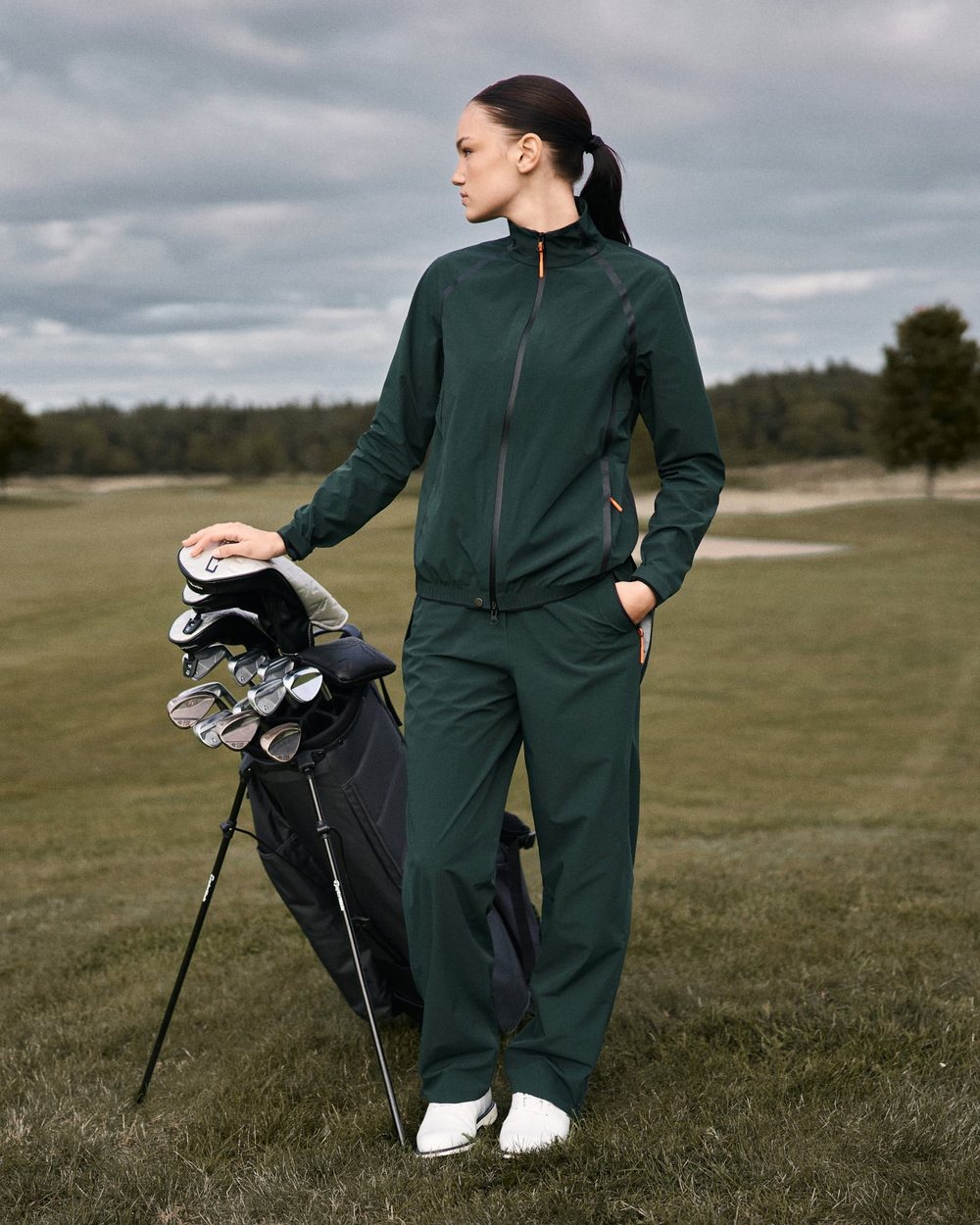 A woman in the green Macade HydroCore rain jacket and trousers beside her golf bag