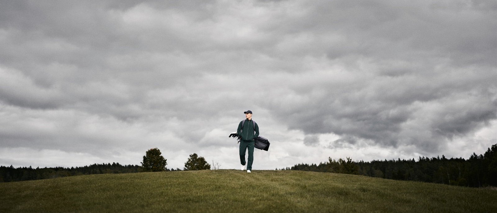 A golfer in a green Macade rain suit walking over a rise with his bag under a heavy grey sky