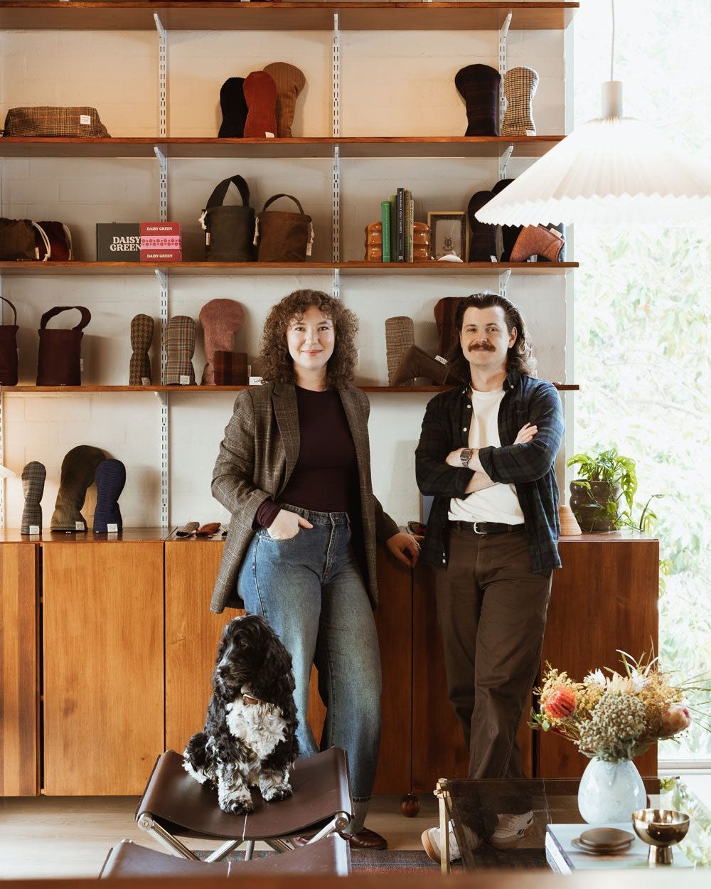 Paige Harding and Adam Brown standing in their studio in front of shelves of headcovers, with their dog