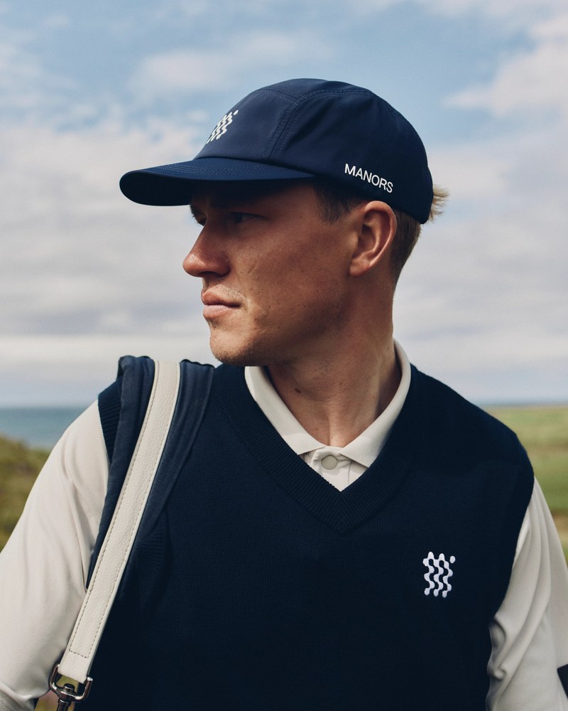 A golfer in a navy Manors sweater vest and cream shirt carrying a bag on a coastal course
