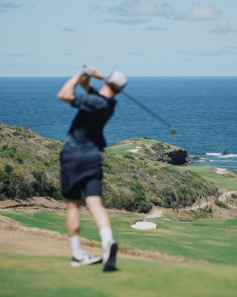 A golfer in a navy Manors shirt swinging on a clifftop tee above the ocean