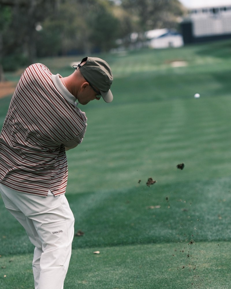 A golfer in a striped Manors polo and olive cap spraying a divot on a fairway