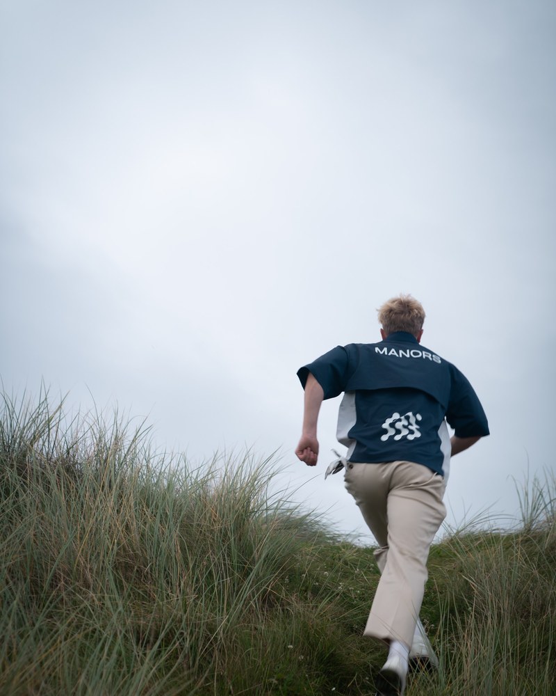 A golfer in a navy Manors shirt running up a grassed dune