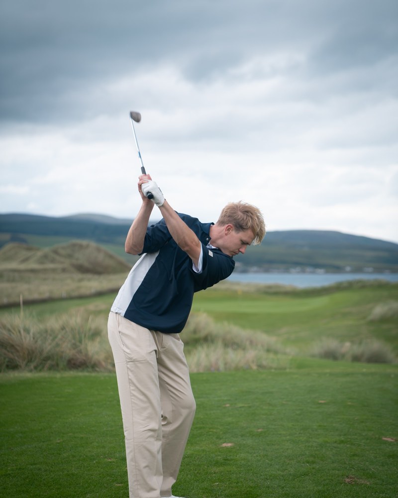 A golfer in a navy Manors polo and stone trousers at the finish of a swing on the links at Machrihanish Dunes
