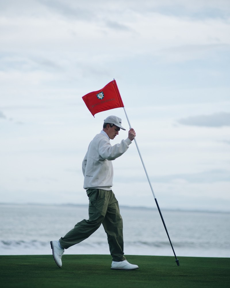 A golfer in a Manors sweater walking off a coastal green holding a red flagstick