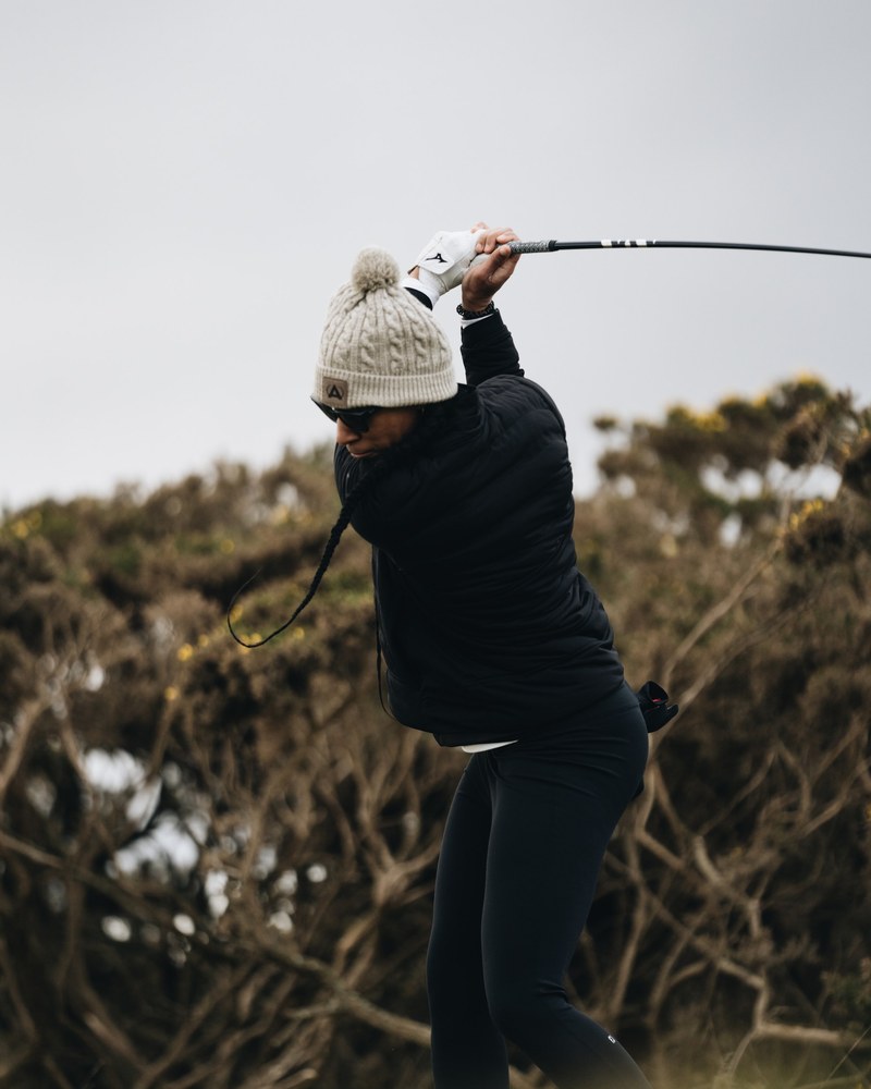 A golfer in a Manors beanie mid-swing on a Scottish links course, gorse in the foreground