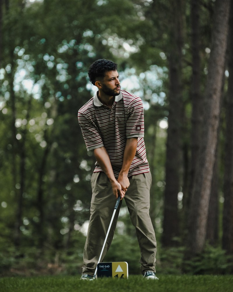 A golfer in a striped Manors polo and olive trousers addressing a ball among pines