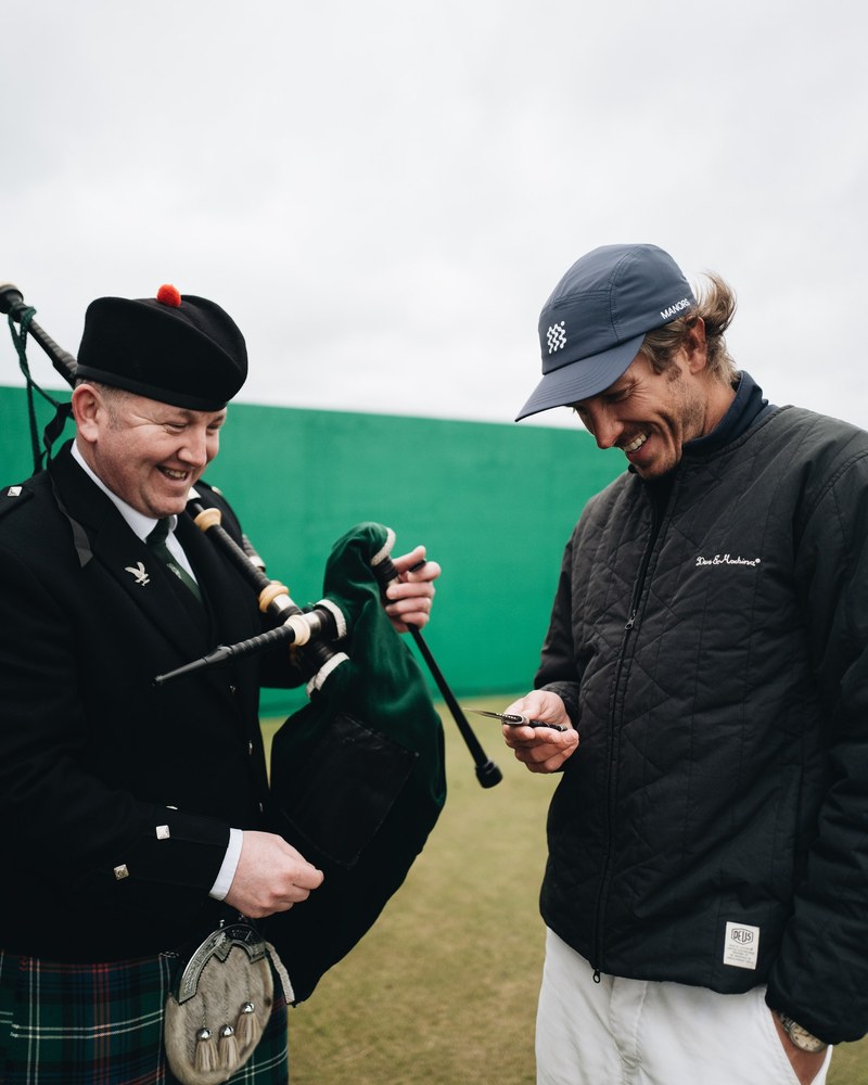 A kilted piper handing a club to a golfer in a Manors jacket at Royal Dornoch