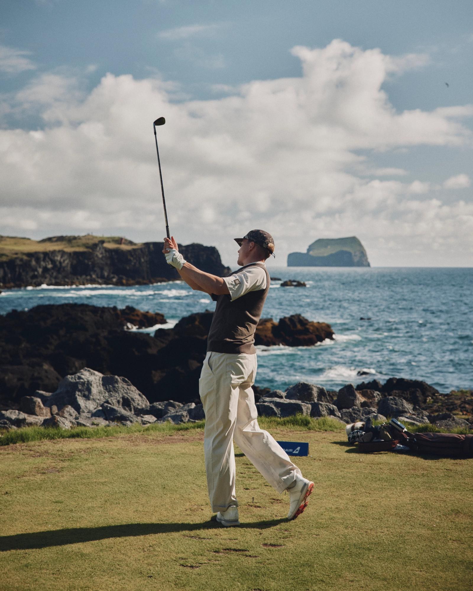A golfer in a Manors polo at the finish of a tee shot on a clifftop fairway with the sea and a rock stack behind