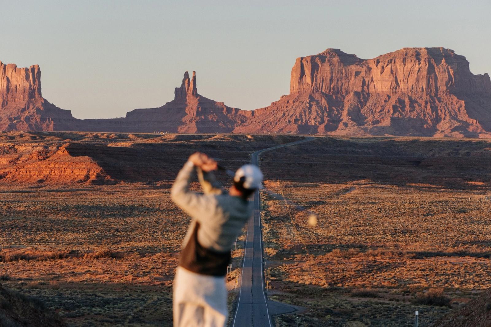 A golfer mid-swing on a desert course with red rock buttes on the skyline