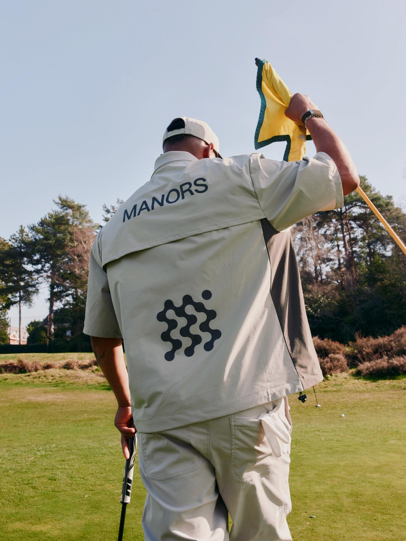 A golfer photographed from behind in a Manors jacket with the MANORS back print, lifting a yellow flag out of the hole