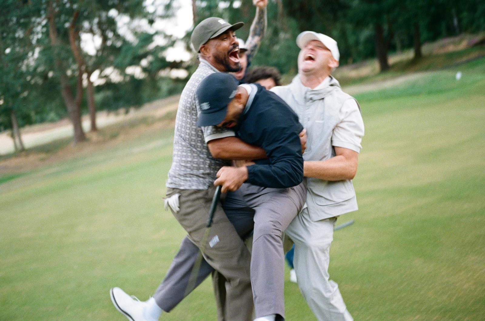 Four golfers piling onto each other laughing on a fairway lined with pines