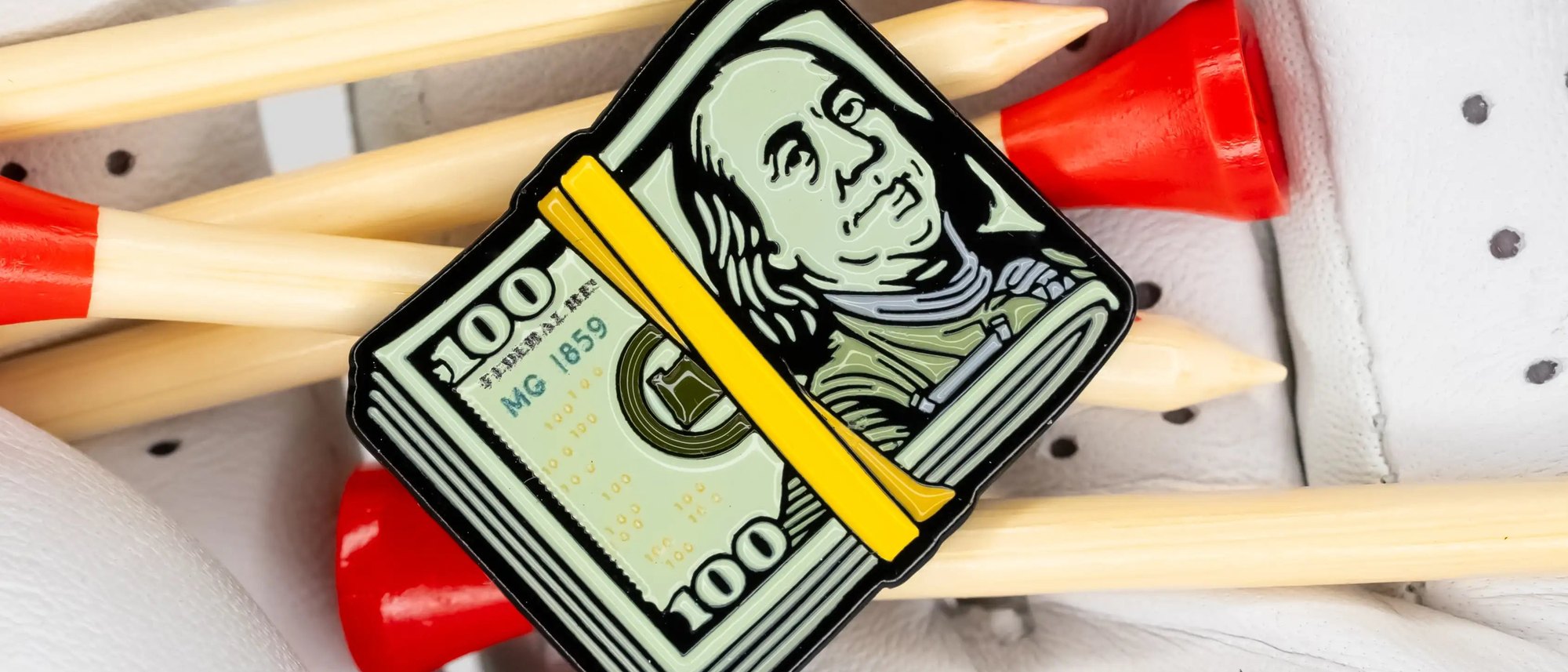 Matchstick Golf’s Cash Stack ball marker, a stack of hundred-dollar bills with a yellow band, on bamboo tees in a white golf glove