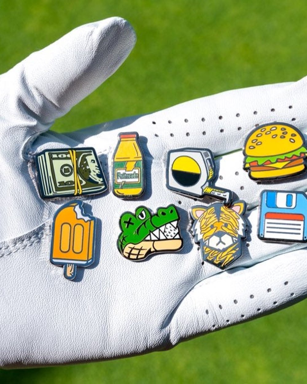 A gloved hand holding eight Matchstick ball markers, including a cash stack, a sports drink bottle and a burger