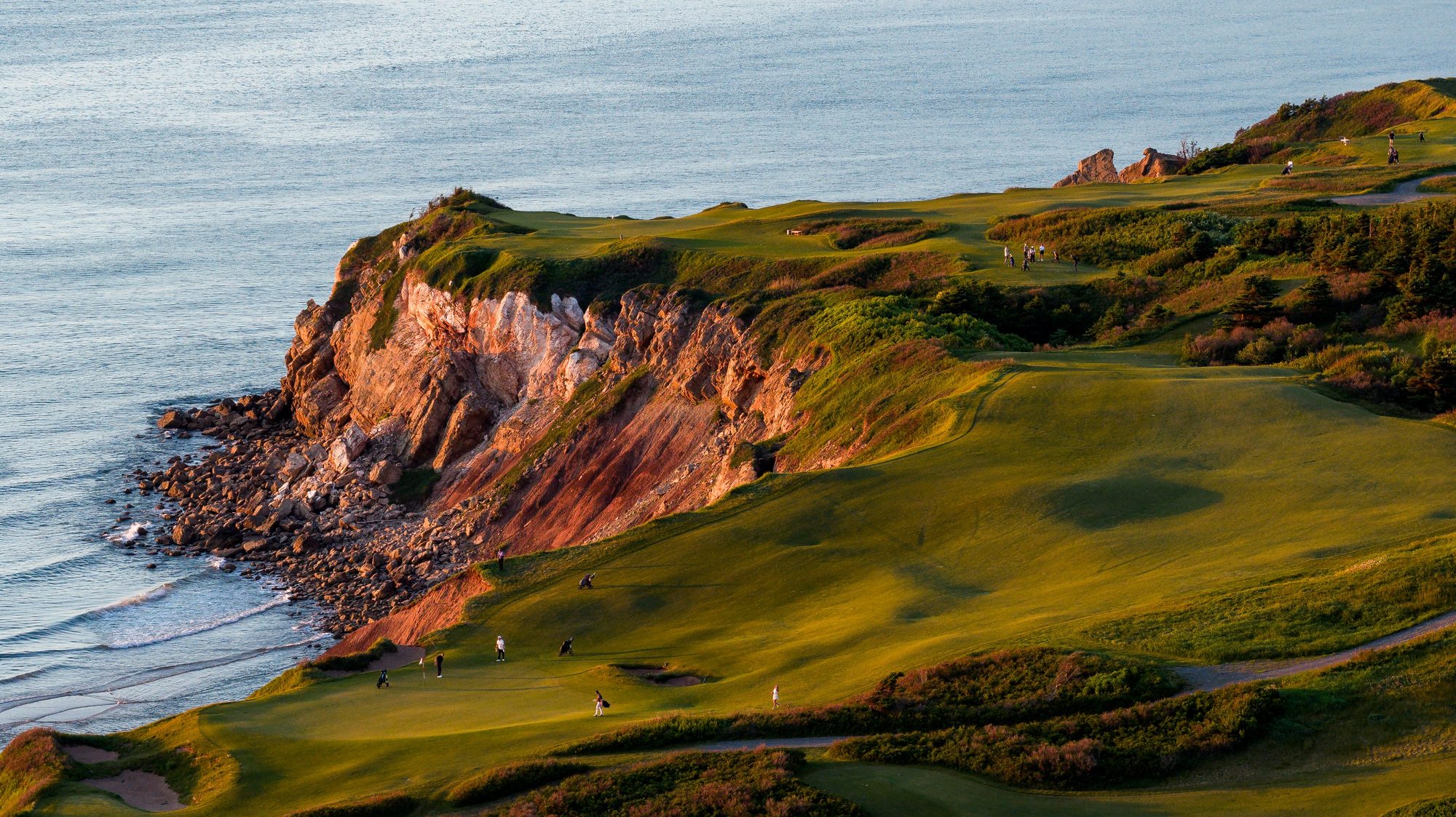 Golfers walking and putting on greens atop a red-rock sea cliff at Cabot Cliffs in golden light