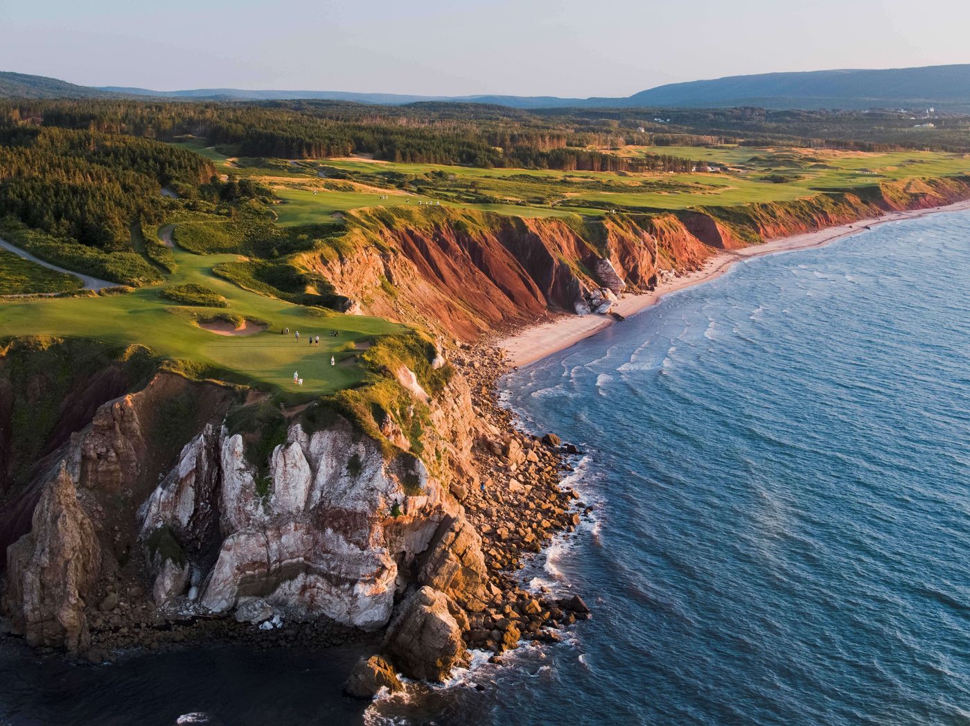 Aerial of greens on top of tall red and white cliffs with a long beach beyond