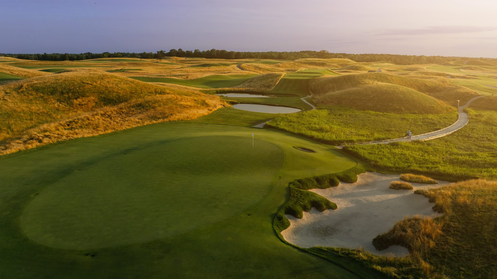 Elevated view over a big Erin Hills green and bunker toward rolling fescue hills at sunset