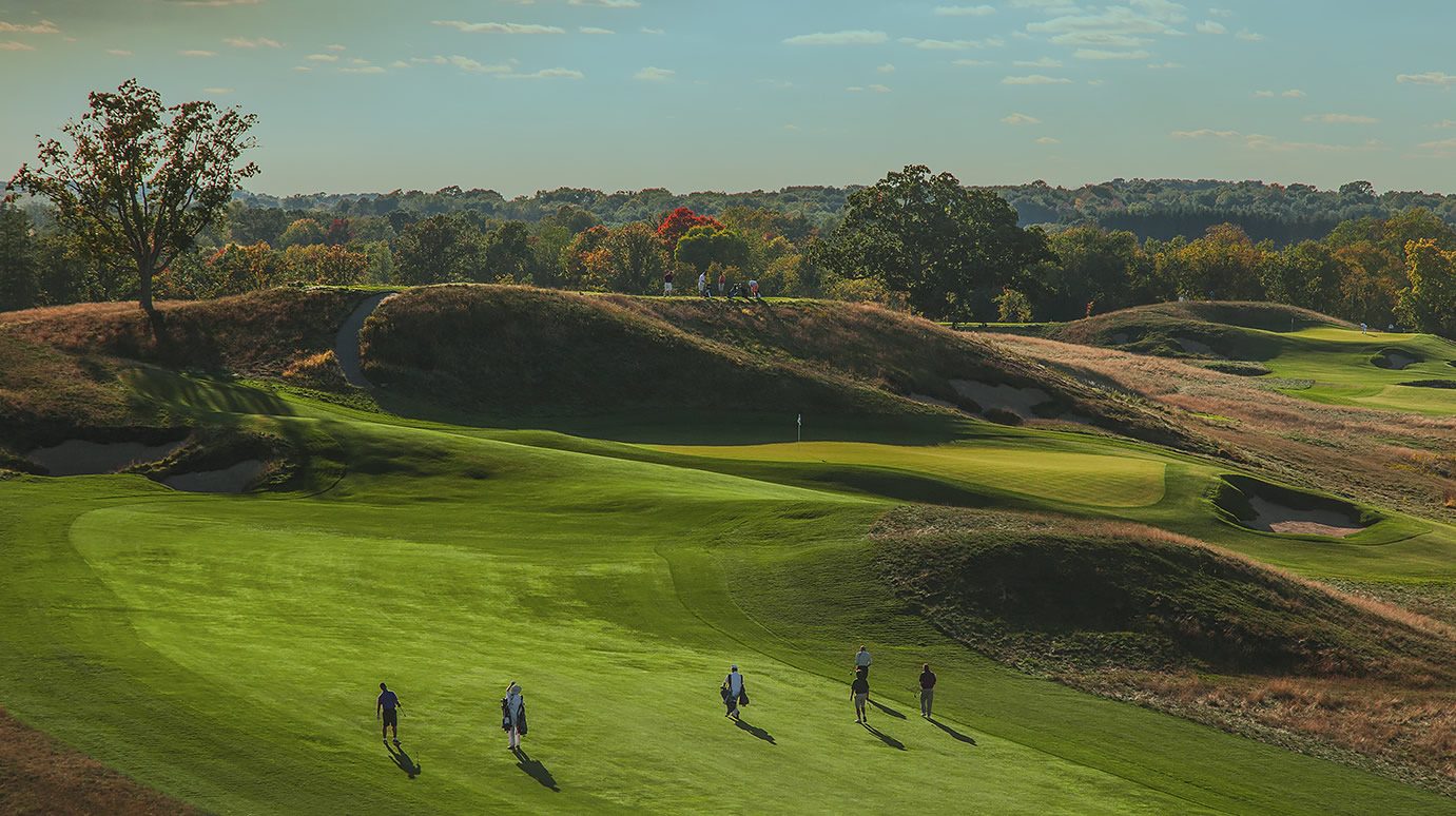 A group of golfers and caddies walking down a sloping Erin Hills fairway in evening light