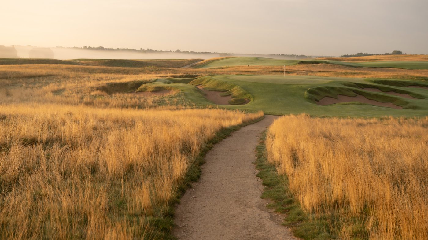 A dirt walking path through waist-high golden fescue toward a bunkered green