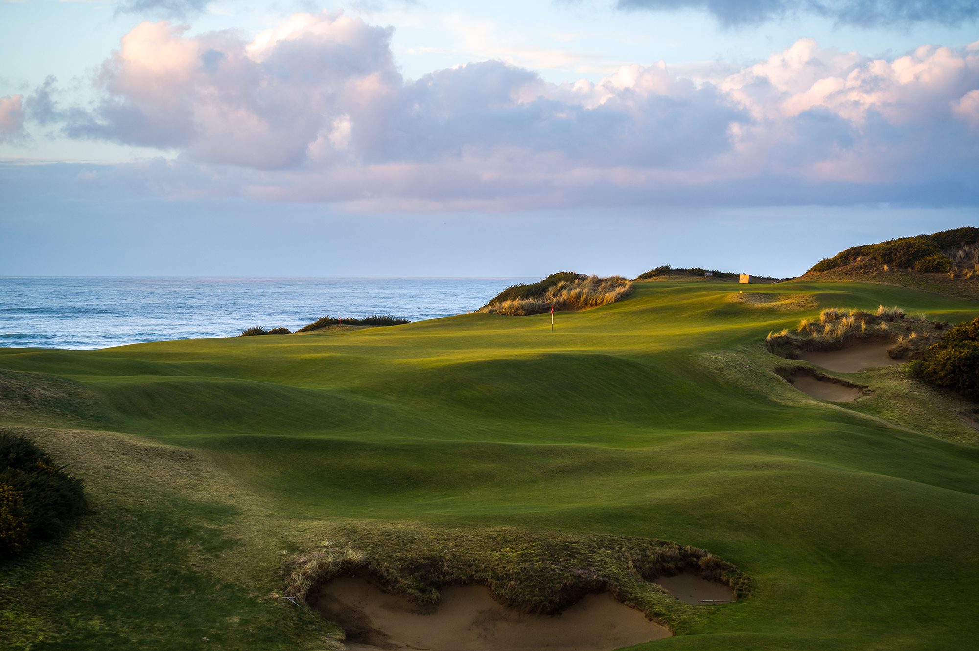 A green perched above the Pacific under big clouds at Old Macdonald, ragged bunker and dune grass in front